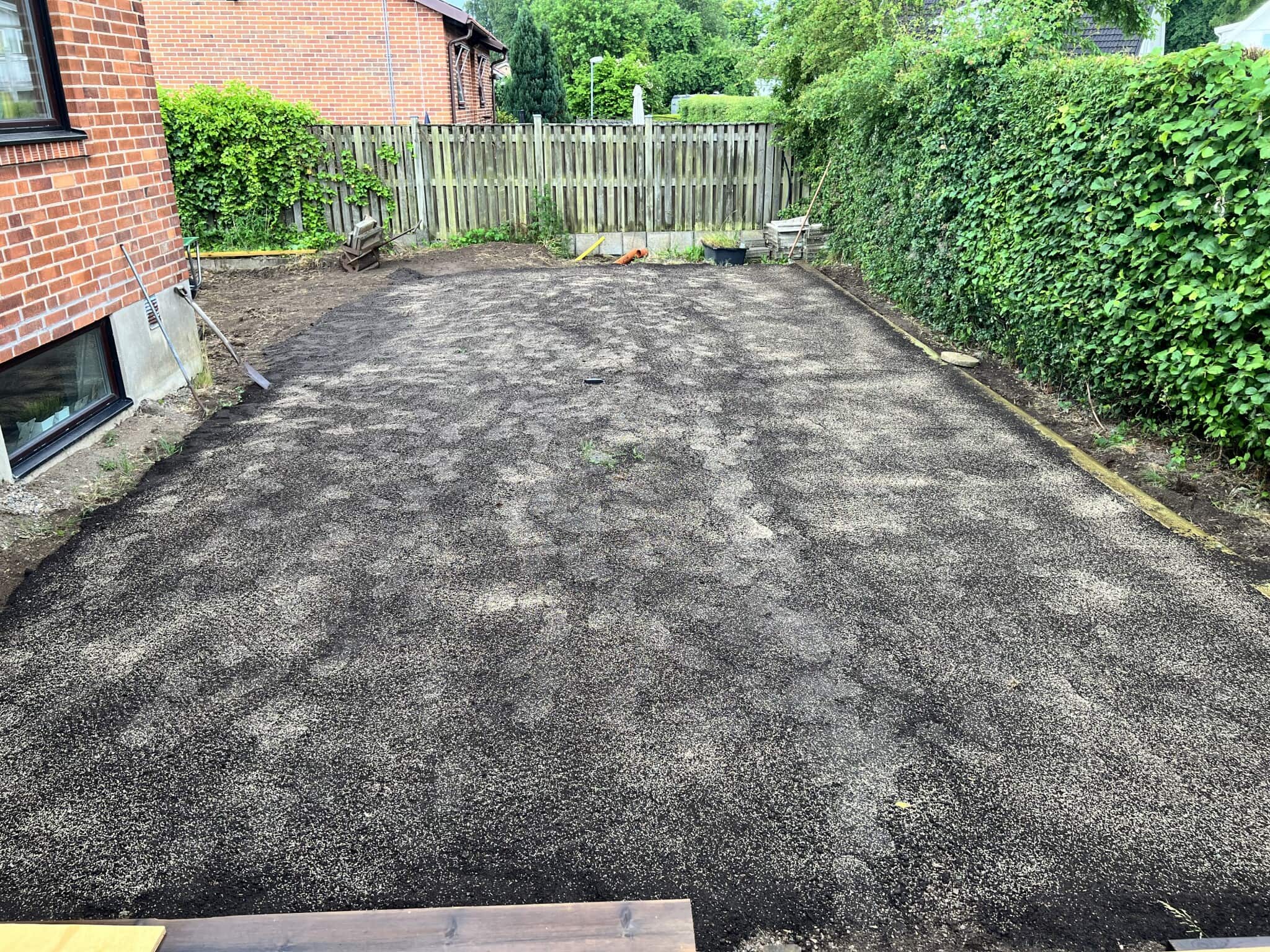 En nyligen grävd och förberedd trädgårdsyta med grus, omgiven av grönska och ett staket, redo för inredning och landskapsdesign.