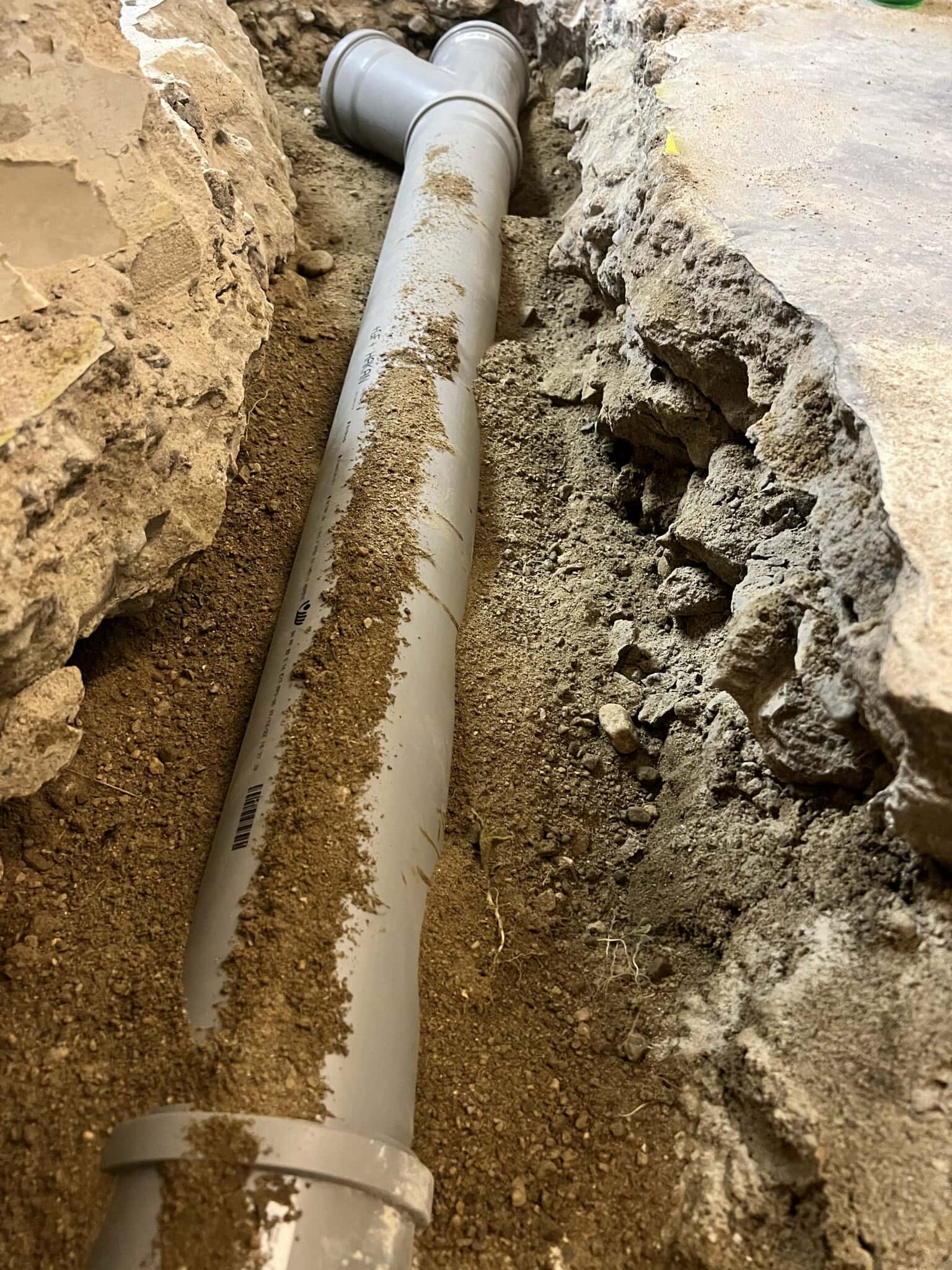 En grå PVC-rörledning ligger i en grävd grop, omgiven av sand och sten, som förbereds för installation.