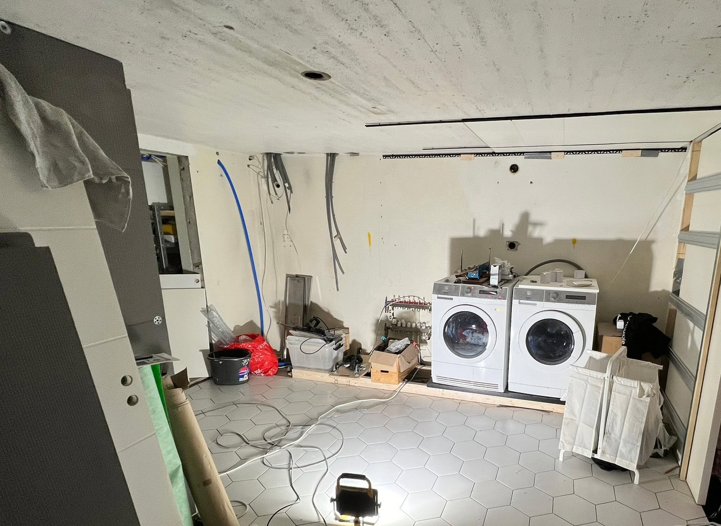 Renovering pågår i tvättstuga med två tvättmaskiner, hexagonalt klinkergolv, lösa kablar och öppna väggar. Känsla av pågående projekt.