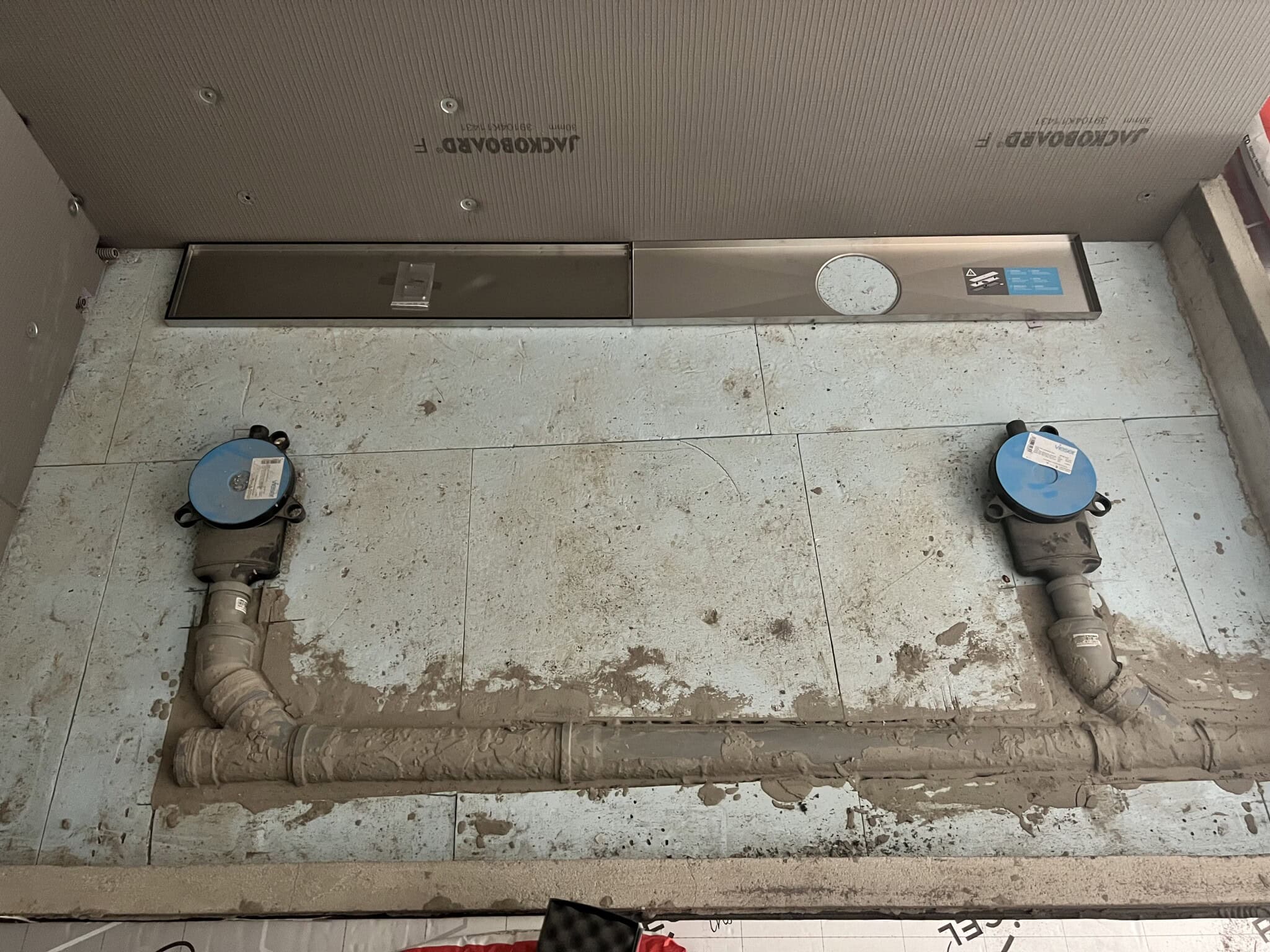 Bilden visar ett pågående badrumsprojekt med synliga rör och installation av VVS-system på en cementbaserad yta.