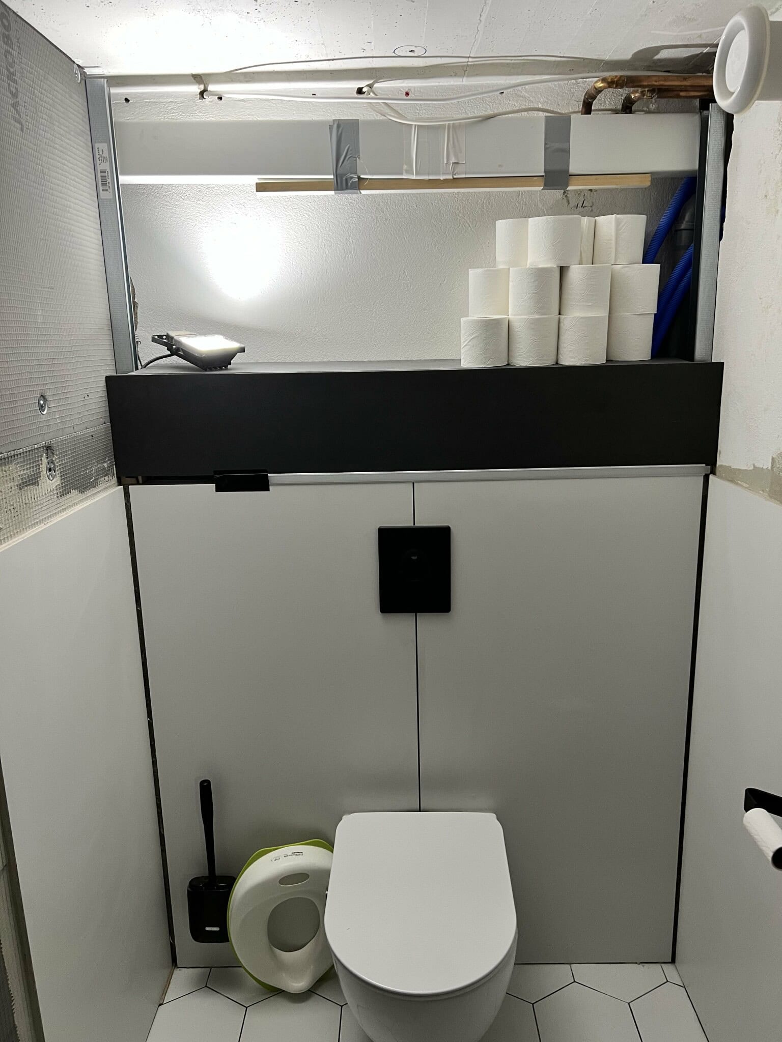 Renoverat badrum med en modern toalett, minimalistisk inredning och en stapel toalettpapper. Belysning och funktionalitet i fokus.