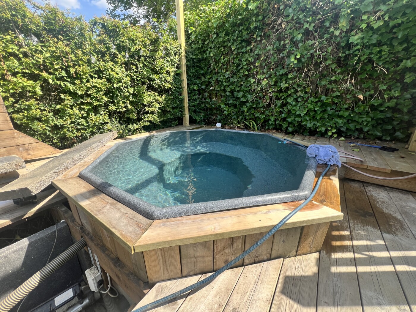 Hexagonformad jacuzzi omgiven av grönska och trädeck.